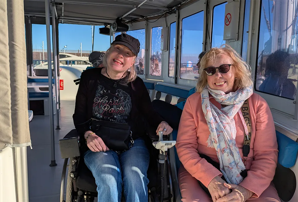 Zwei Frauen sitzen lächelnd auf einem Ausflugsboot, eine davon im Rollstuhl – barrierefreie Bootsfahrt, Inklusion und gemeinsames Ferienerlebnis auf dem Wasser.