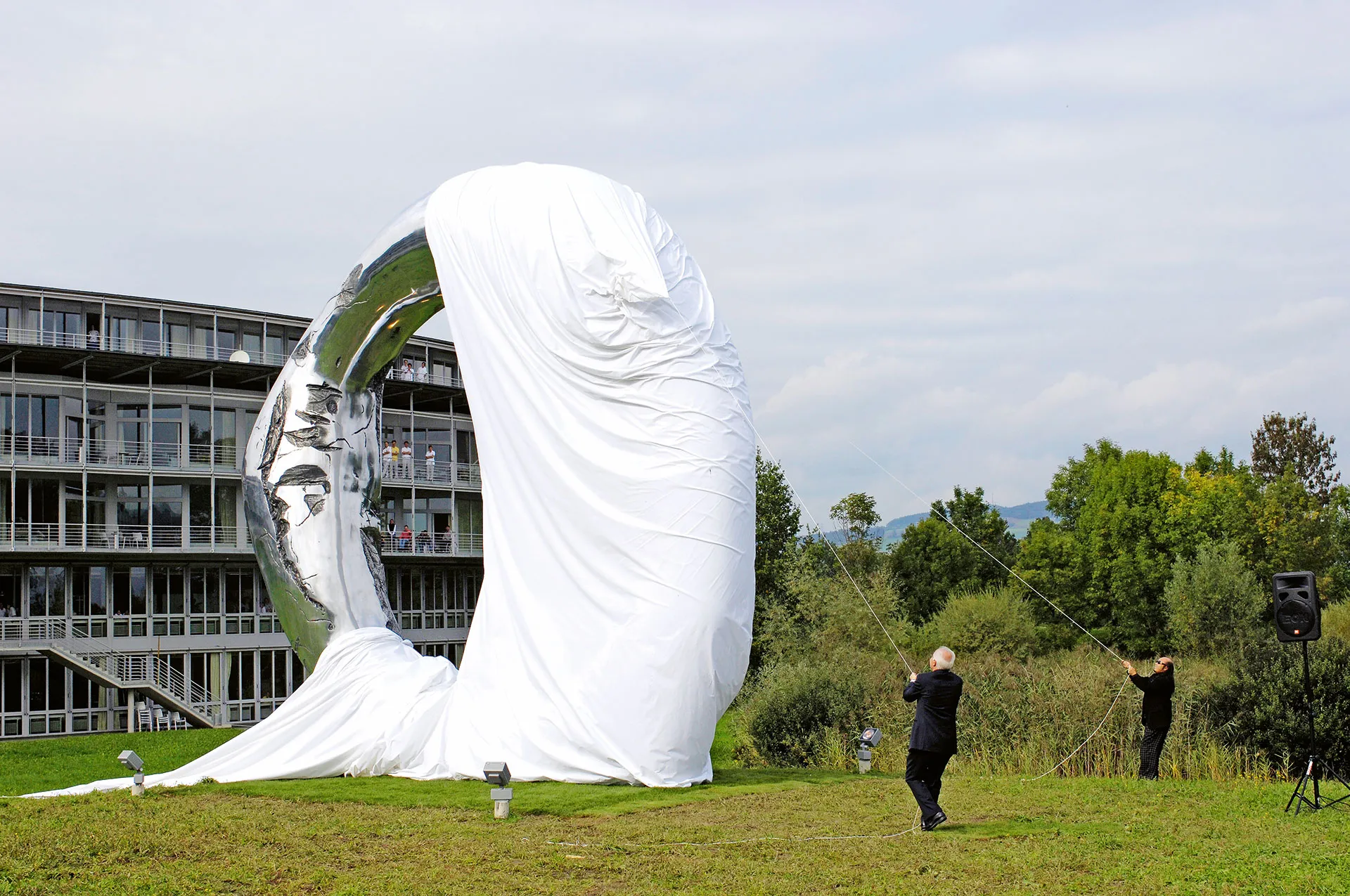 A large white sculptural form, resembling a giant snail shell, stands in a grassy field in front of a modern glass building.