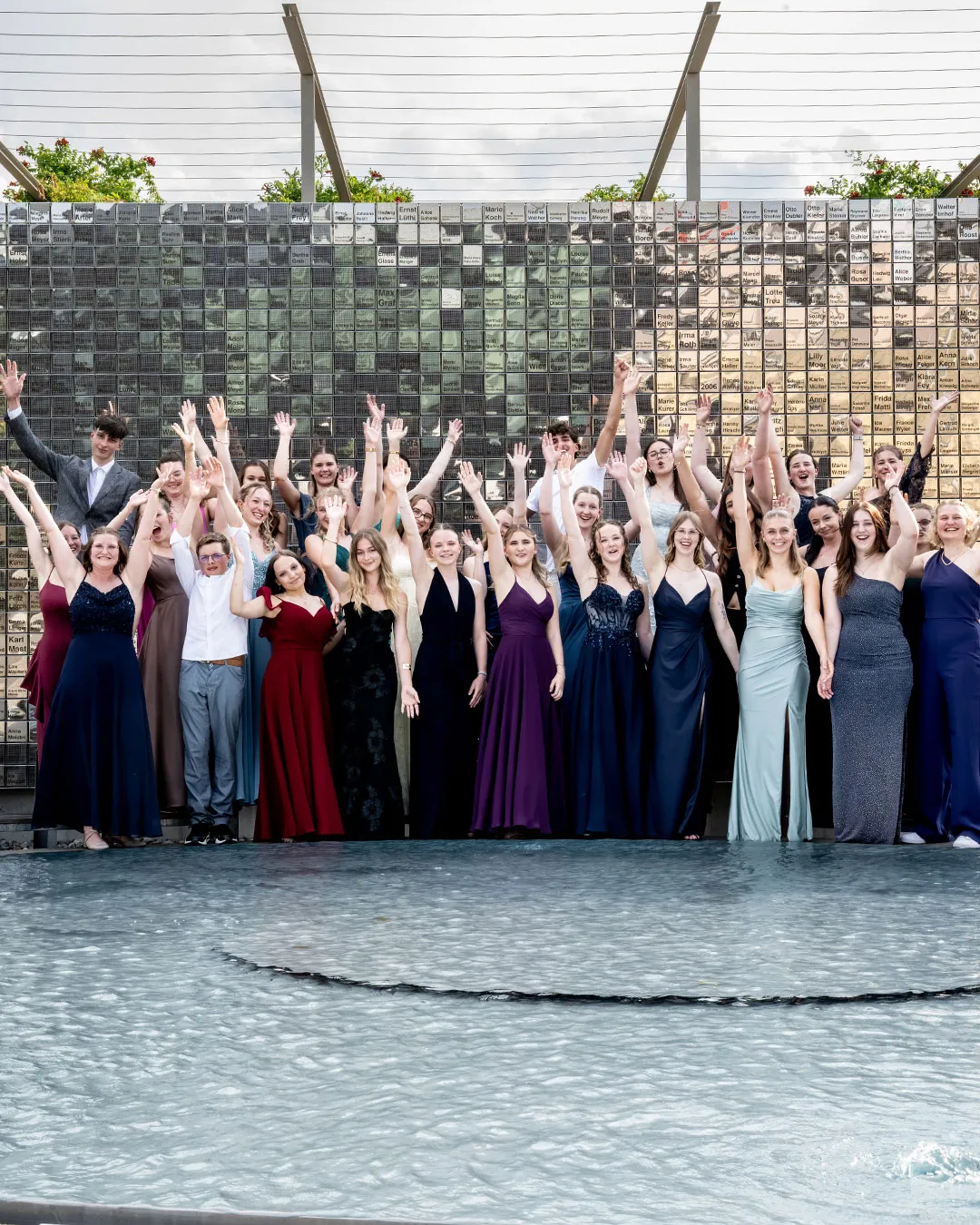 A large group of people in coordinated colorful dresses standing and posing together outdoors in front of a stone wall and water feature.