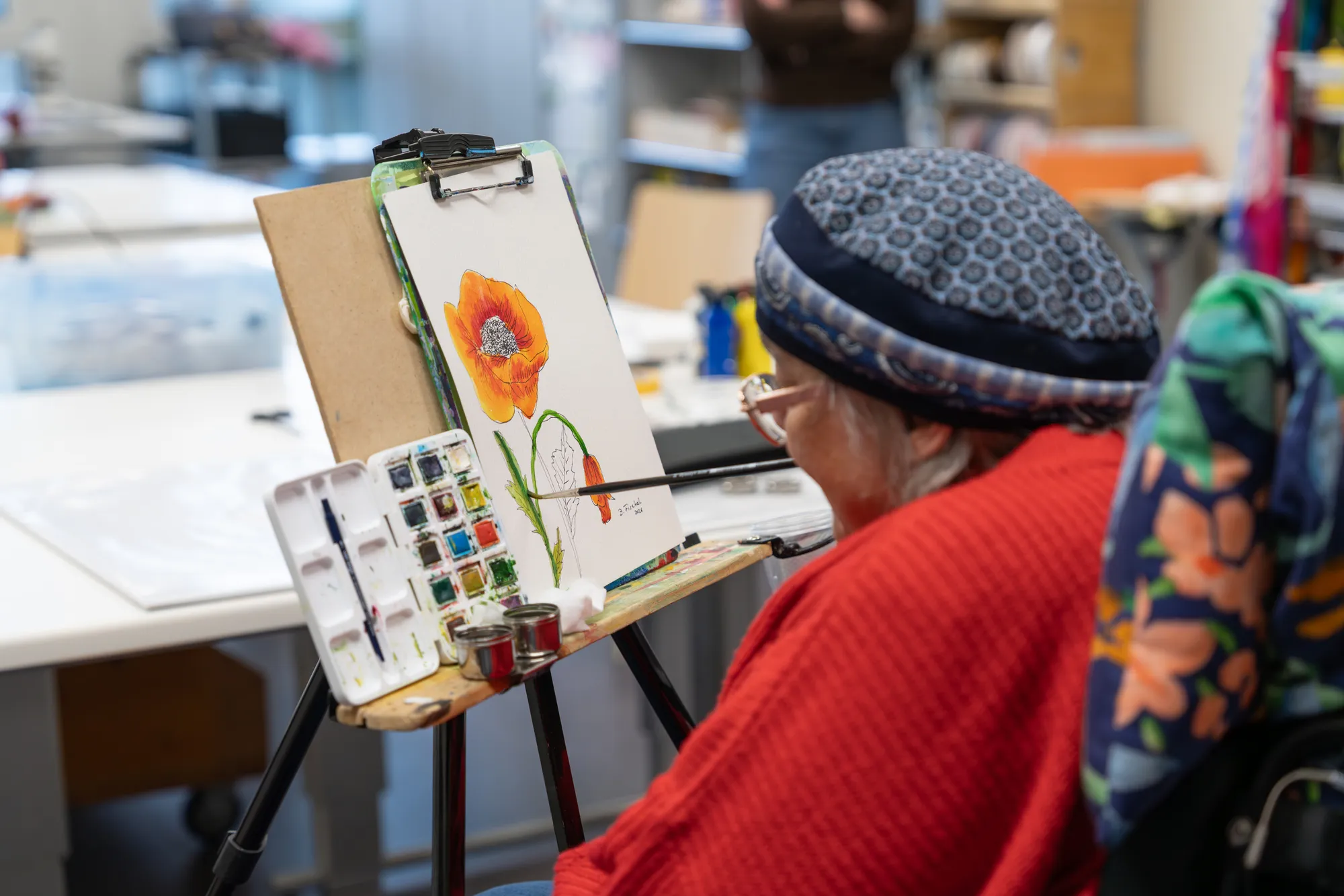 A gray-haired man wearing a red sweater and cap stands at an easel, painting a landscape with a paintbrush.