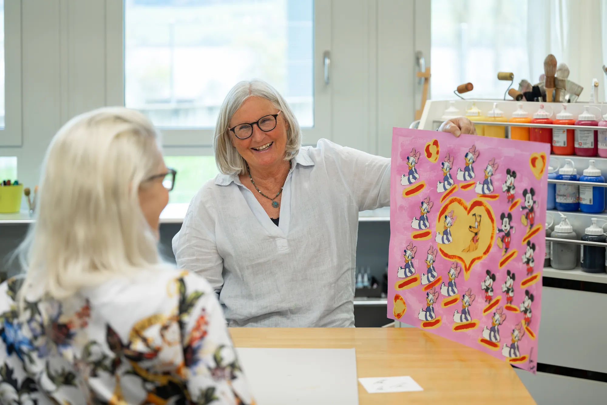Two older women conversing in a colorful, well-lit classroom setting.