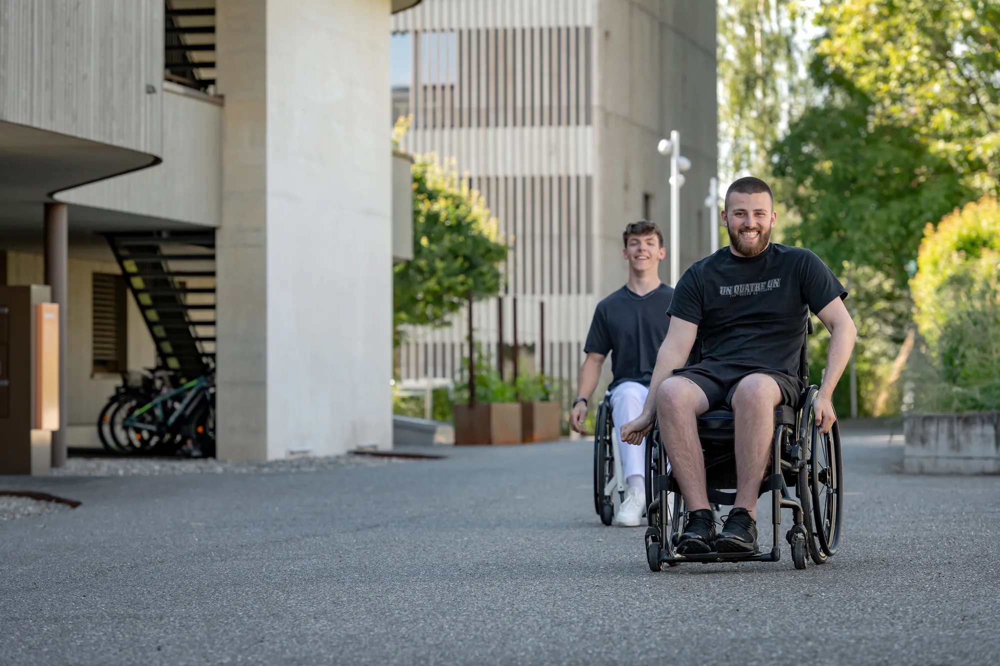 A man in a wheelchair is smiling and looking at the camera as he goes down a sidewalk on a sunny day.