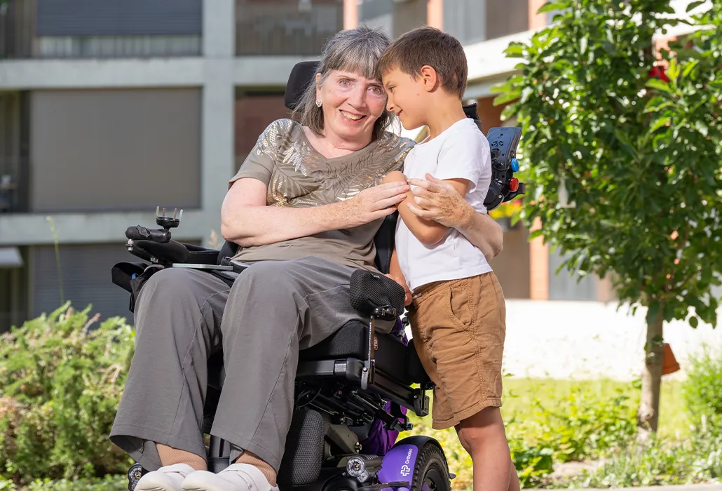 A smiling woman in a wheelchair embracing a young boy outdoors.