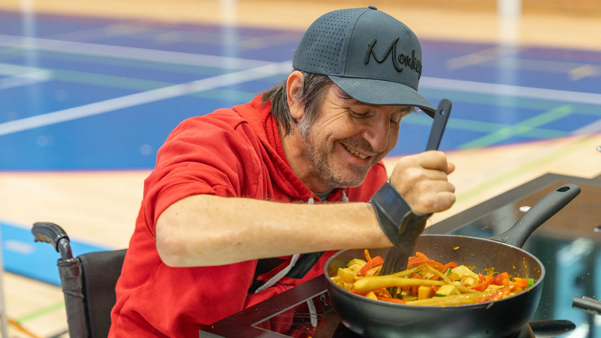 A bearded man in a red shirt and baseball cap concentrates while eating food from a bowl on his lap, seated in an outdoor setting.