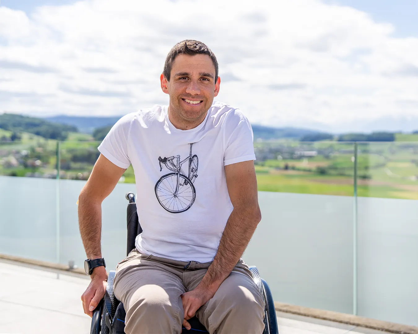 A smiling man in a white t-shirt and wheelchair overlooking a scenic landscape.