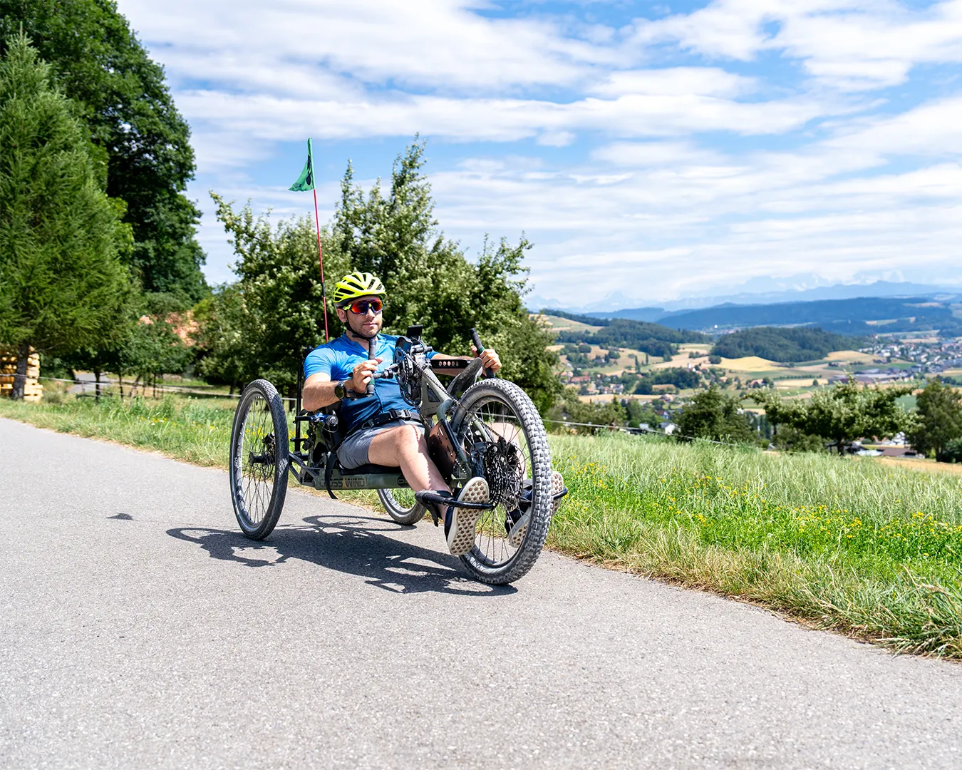 A cyclist rides a recumbent trike on a rural road surrounded by green trees and hills.