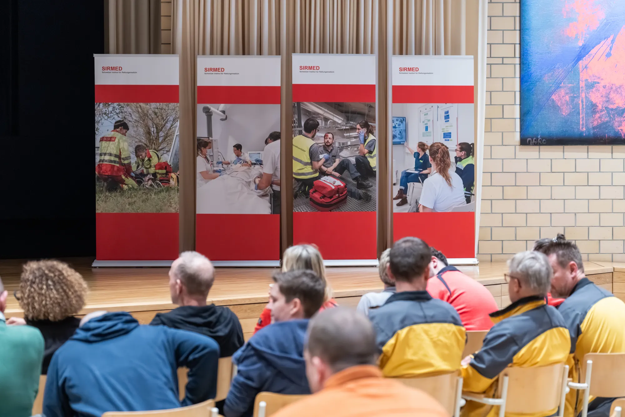 A small crowd of people sitting in a lecture hall and watching a presentation on large screens.