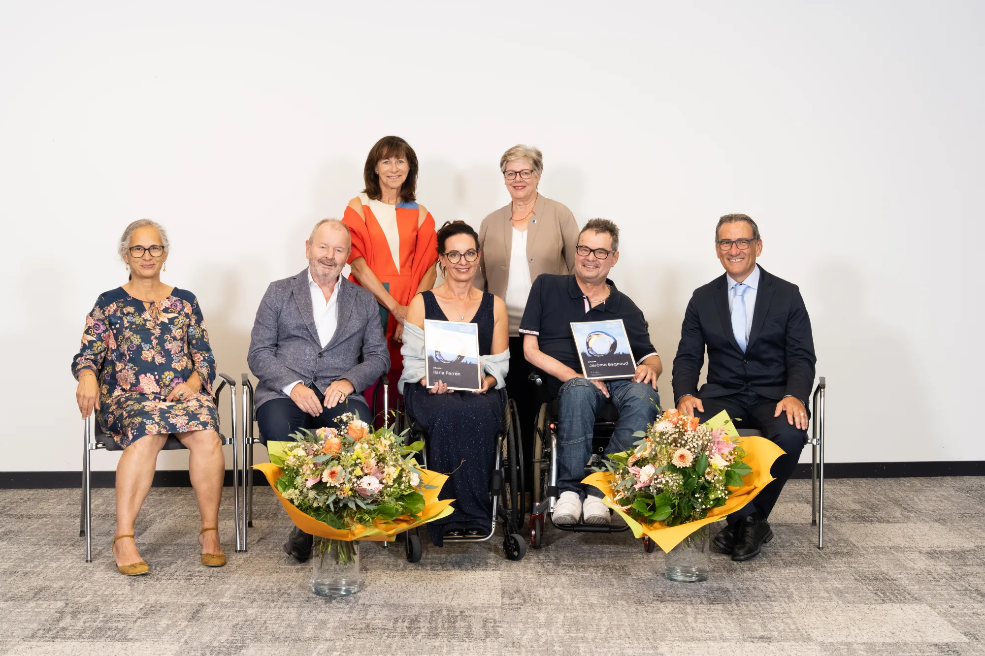 A group portrait of six people, including two women and four men, sitting and standing in front of a plain white wall. They are all smiling and holding bouquets of flowers.