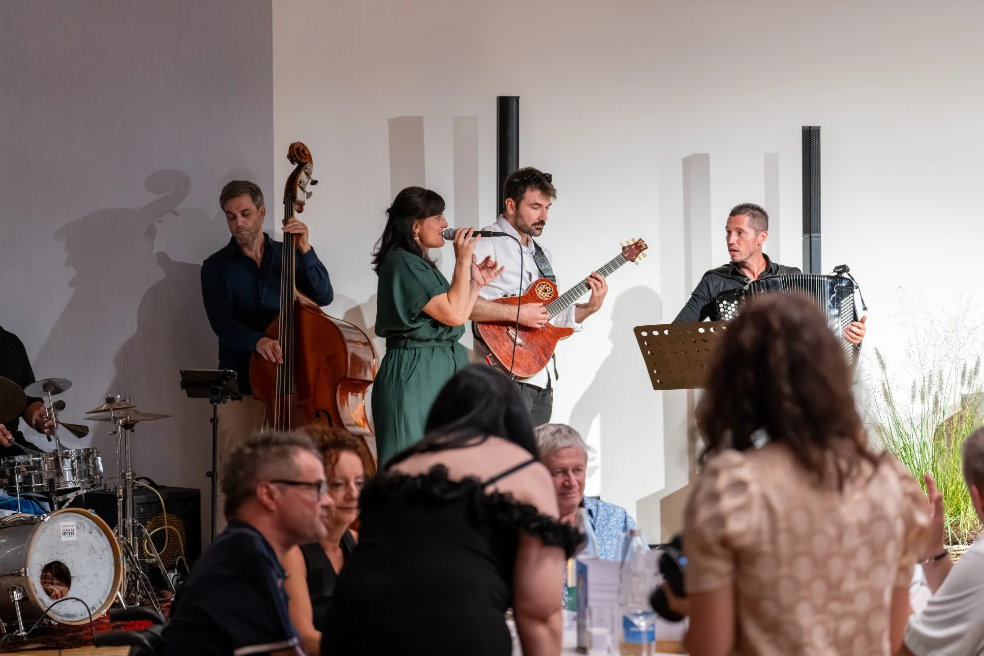 A group of musicians performing on a small stage, with electric and acoustic guitars, drums, and a bass player.