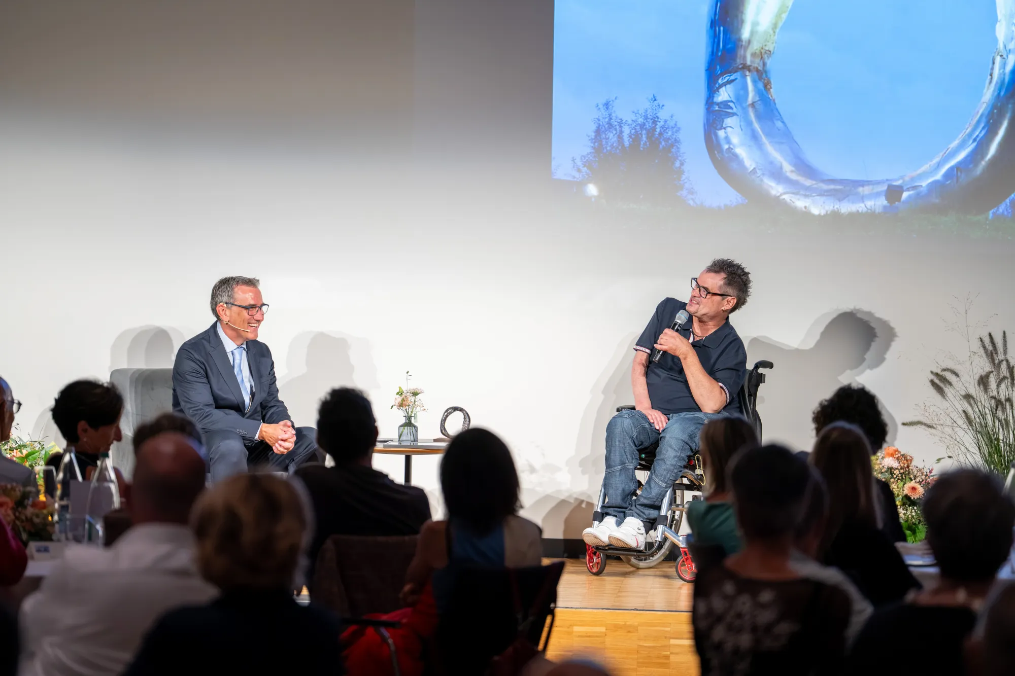 A large crowd of people attending an event, with two men on a stage addressing the audience.