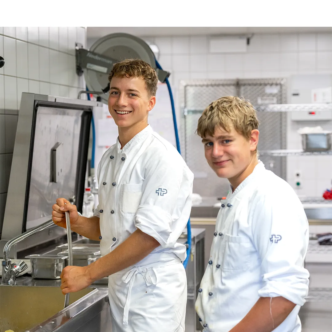 Two young chefs in a kitchen setting, wearing white uniforms and chef hats.