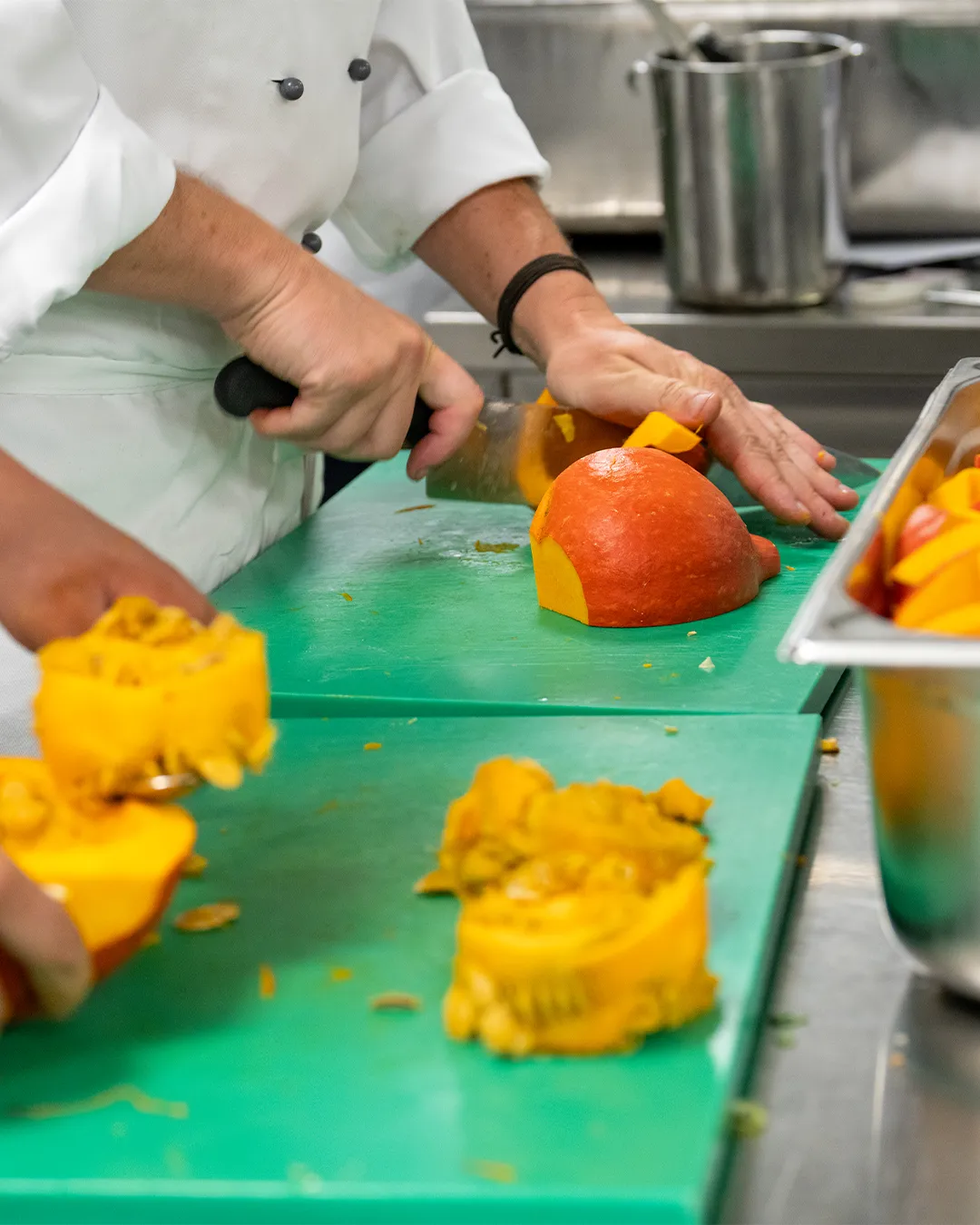 A chef's hands chopping pumkin and yellow bell peppers on a green cutting board in a kitchen.
