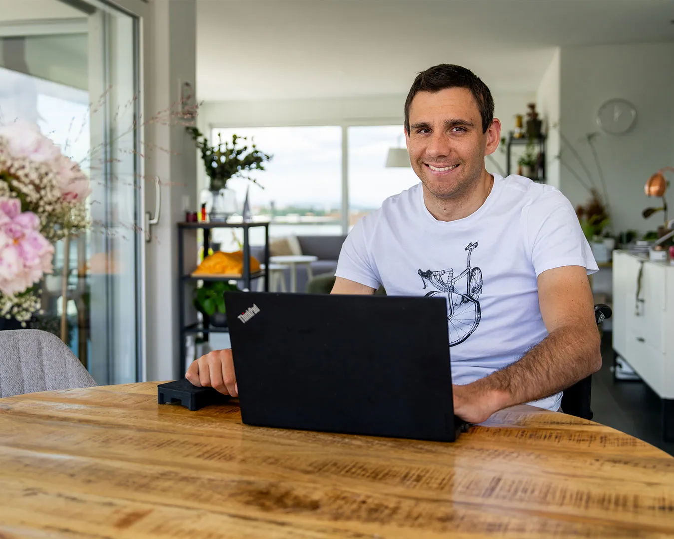 A smiling man sitting at a table with a laptop, surrounded by house plants.