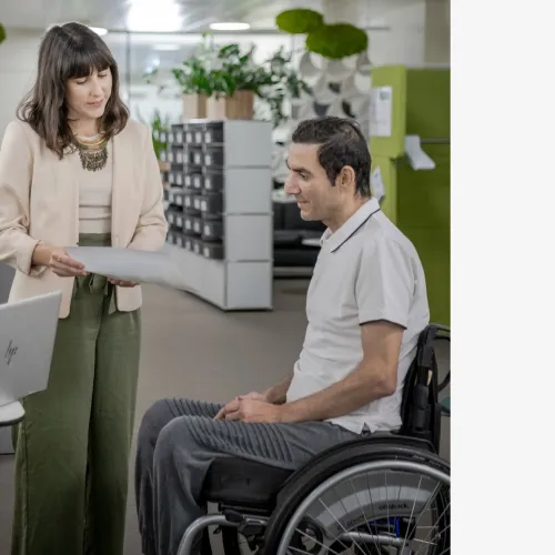 In einem modernen Büro sind zwei Personen in einem Gespräch. Die Frau links, Selina Infanger trägt eine helle Jacke, ein helles Oberteil und grüne Hosen. Sie steht, hält ein Dokument in der Hand und liest etwas darauf vor. Der Mann rechts sitzt in einem Rollstuhl und trägt ein weißes T-Shirt und graue Hosen. Vor ihnen auf einem kleinen Tisch steht ein aufgeklappter Laptop. Der Hintergrund zeigt eine offene Büroeinrichtung mit Pflanzen und modernen Dekorationselementen wie hängenden grünen Moosobjekten.