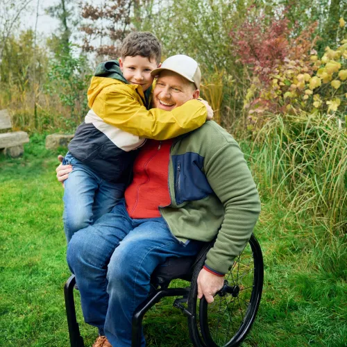Heiri sitzt in seinem Rollstuhl auf einer Wiese und sein Sohn klettert ihm halbwegs auf den Schoss und umarmt seinen Vater ganz fest. Beide lächeln.