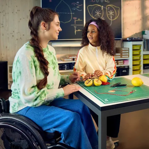 Chiara Schlatter sitzt im Rollstuhl an einem Tisch in einem Schulzimmer zusammen mit einer Schülerin und zeigt ihr, wie man mit Zitronen eine Lampe zum Leuchten bringen kann. Beide schauen sich an und lächeln.