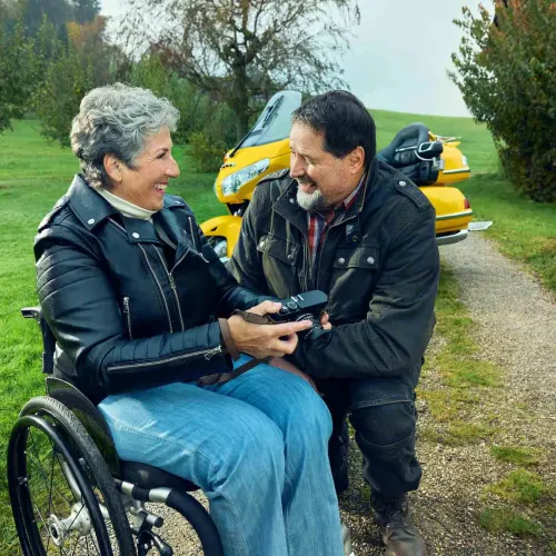 Rosa Zaugg sitzt im Rollstuhl auf einem Kiesweg, hält eine Kamera in der Hand und lächelt ihren Partner Lukas an.