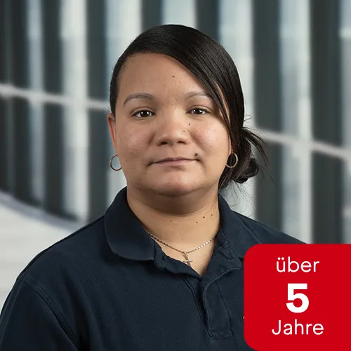 Porträt einer lächelnden Frau mit dunklen Haaren in Arbeitskleidung vor einem Bürogebäude.