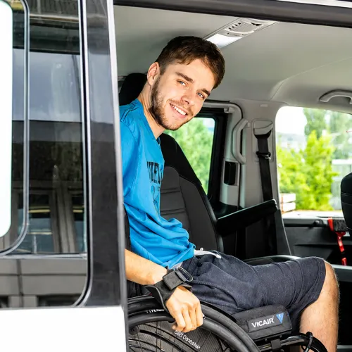 Smiling man in blue shirt seated inside vehicle, holding steering wheel.