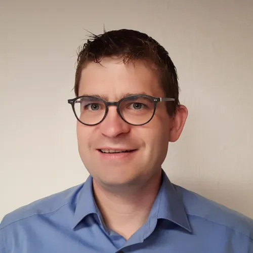 A smiling man with glasses wearing a light blue shirt.