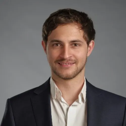 A cheerful man with brown hair and a slight smile, wearing a white shirt and dark suit jacket.