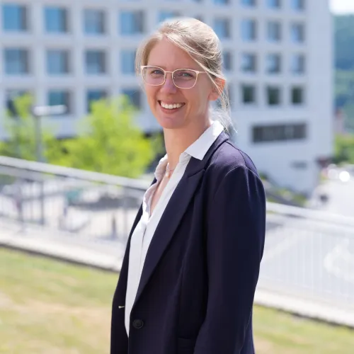 A smiling middle-aged woman with blonde hair wearing glasses and a dark jacket stands outdoors in front of a modern building.
