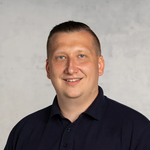 Ein lächelnder Mann mit kurzen dunklen Haaren in einem blauen Hemd vor einem grauen Hintergrund.