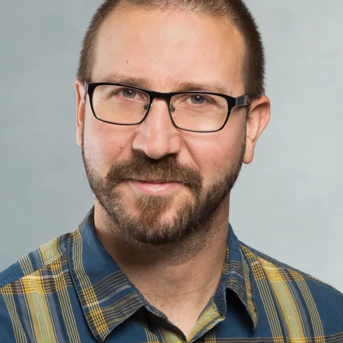 A bearded man wearing glasses and a plaid shirt, looking directly at the camera with a serious expression.