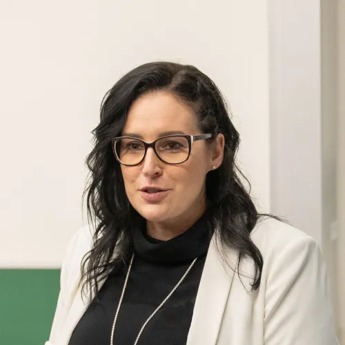 A woman with curly dark hair wearing glasses and a white jacket, looking at the camera with a serious expression.