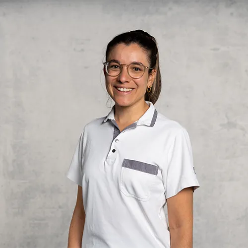 A smiling young man wearing glasses and a white polo shirt stands against a gray wall.