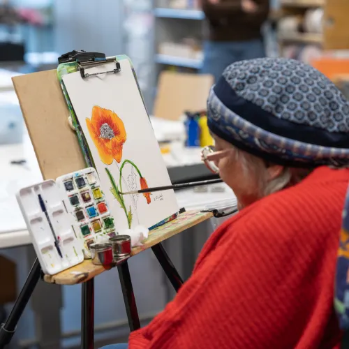 A gray-haired man wearing a red sweater and cap stands at an easel, painting a landscape with a paintbrush.