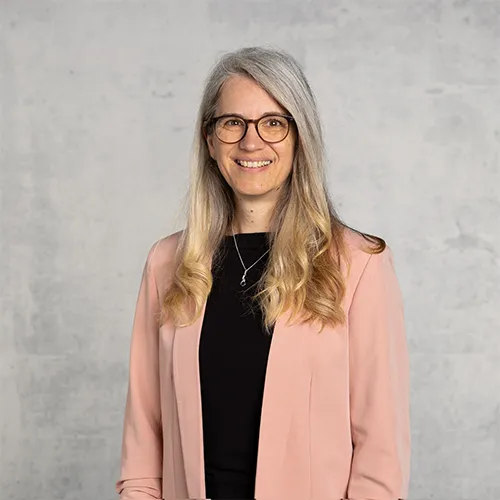 Smiling woman with long grey hair wearing glasses and a pink jacket against a plain background.