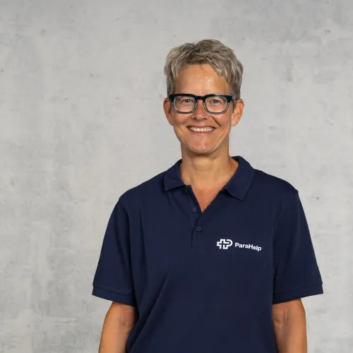 Porträt einer Frau mit kurzen grauen Haaren und Brille, die ein dunkelblaues Hemd mit dem Logo eines Unternehmens trägt.