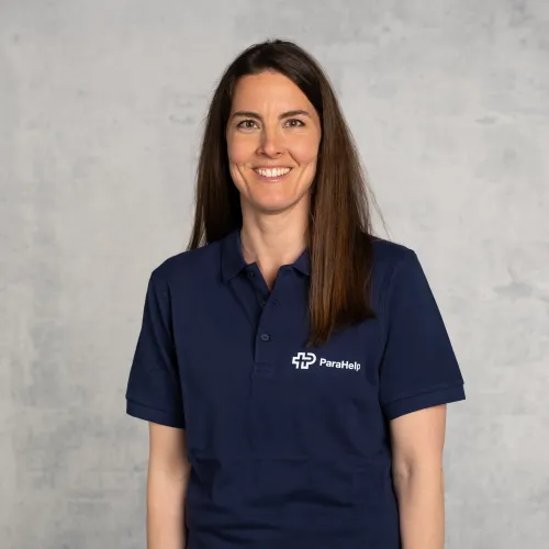 Eine Fotografie einer lächelnden Frau mit langem, braunem Haar, die eine marinelaue Poloshirt mit Logo trägt.