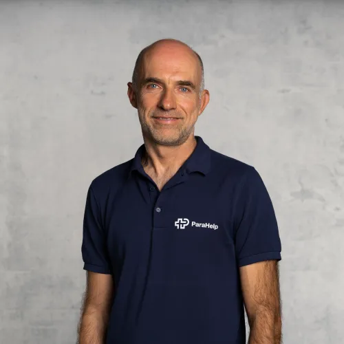 Ein männliches Porträt vor einem grauen Hintergrund. Der Mann trägt ein dunkelblaues Poloshirt mit einem Logo.