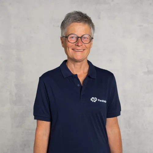 Eine lächelnde Frau mittleren Alters mit grauem Haar und Brille, die eine marineblaue Polo-Bluse mit einem Logo trägt.