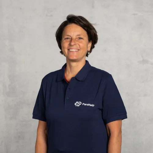 Eine Frau mit kurzen dunklen Haaren, die lächelnd in die Kamera schaut und ein dunkelblaues Poloshirt mit einem Logo trägt.