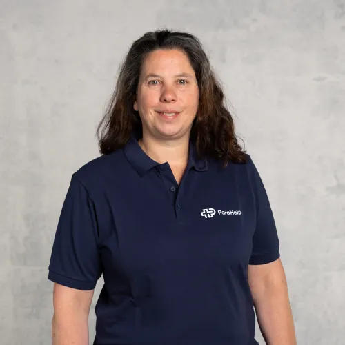 Ein freundliches Porträt einer Frau mit langen braunen Haaren, die einen dunkelblauen Poloshirt mit einem weissen Logo trägt.