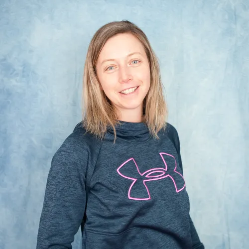 A smiling woman with long blonde hair wearing a navy blue sweatshirt with the Under Armour logo visible.