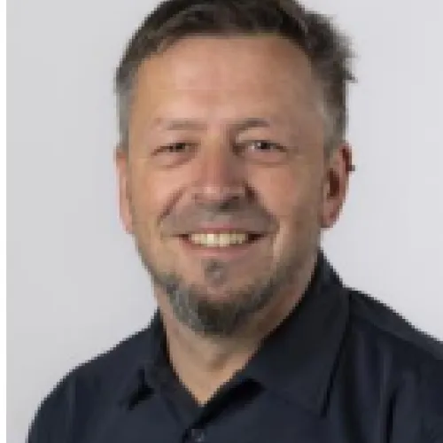 Smiling middle-aged man with short dark hair and beard, wearing a blue collared shirt.