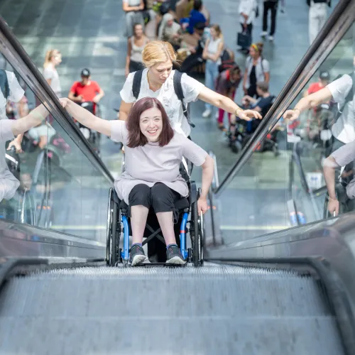 Una donna in sedia a rotelle sale su una scala mobile, seguita da un’assistente.